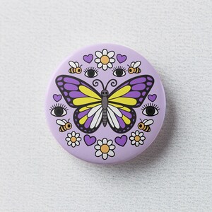 K&ouml;nnte beinhalten: Ein runder, lila Knopf mit einem Schmetterling mit gelben und lila Fl&uuml;geln. Der Knopf ist mit Bienen, Blumen, Herzen und Augen verziert. Der Schmetterling ist das zentrale Designelement, umgeben von anderen skurrilen Illustrationen.