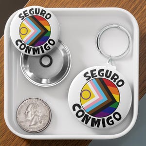 May include: A white button with the text "Seguro Conmigo" in black lettering and a rainbow flag with a yellow circle in the center. The button is on a white surface with a silver button back and a quarter.