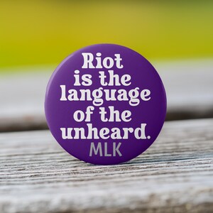 May include: A round, purple button with white text that reads "Riot is the language of the unheard. MLK". The button is set against a blurred background of green and yellow.