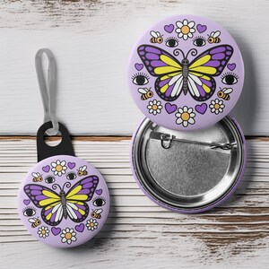 K&ouml;nnte beinhalten: Ein Set aus lavendelfarbenen Ansteckbuttons und einem Schl&uuml;sselanh&auml;nger mit Schmetterlingsdesign. Der Schmetterling hat lila und gelbe Fl&uuml;gel, umgeben von Bienen, G&auml;nsebl&uuml;mchen, Herzen und Augen. Der Schl&uuml;sselanh&auml;nger hat eine schwarze Schlaufe.