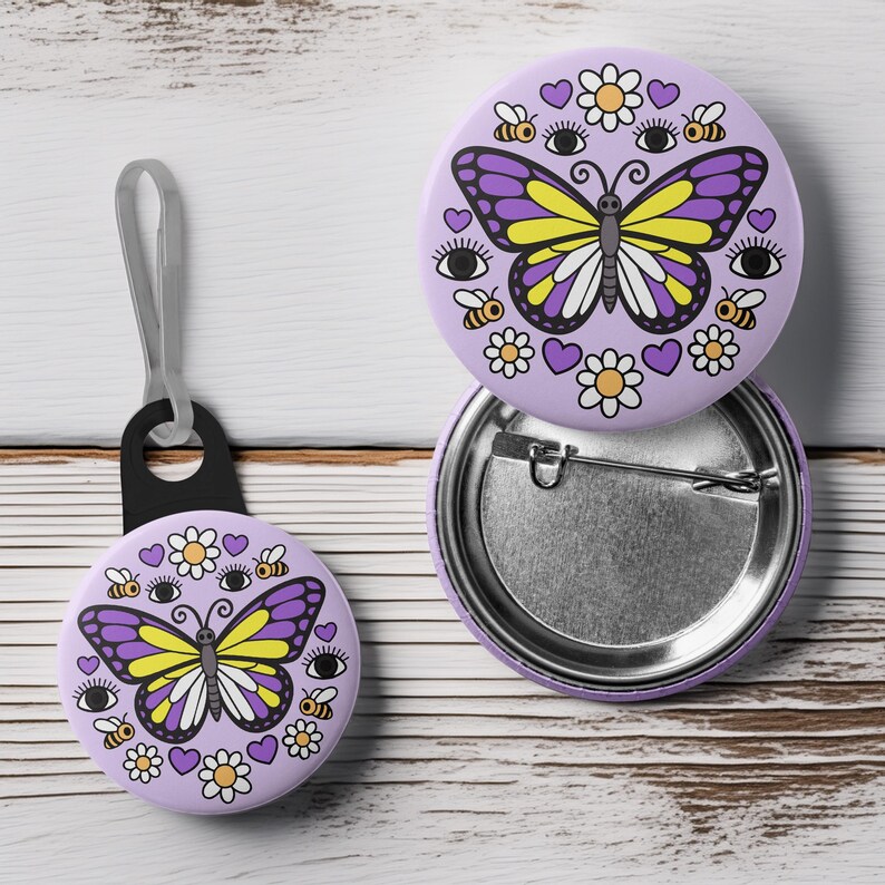 K&ouml;nnte beinhalten: Ein Set aus lavendelfarbenen Ansteckbuttons und einem Schl&uuml;sselanh&auml;nger mit Schmetterlingsdesign. Der Schmetterling hat lila und gelbe Fl&uuml;gel, umgeben von Bienen, G&auml;nsebl&uuml;mchen, Herzen und Augen. Der Schl&uuml;sselanh&auml;nger hat eine schwarze Schlaufe.