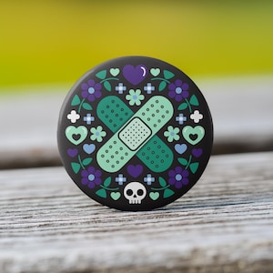 May include: A round, black button with a decorative design. The design features a green and teal bandage, surrounded by purple flowers, hearts, and a small skull. The button is set against a blurred green and wood background.