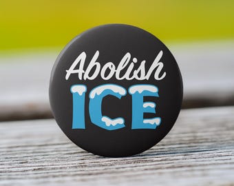 May include: A circular black pin-back button featuring the white text Abolish in a cursive script font above the word ICE in a bold blue block font, with white snow-like accents layered on top of the blue letters.