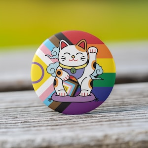 May include: A round button featuring a white, orange, and black Maneki-neko cat with a raised paw. The button has a rainbow background and includes the colors of the Progress Pride flag. The cat is sitting on a purple cushion.