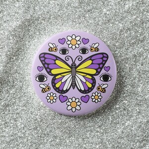 K&ouml;nnte beinhalten: Ein runder, lavendelfarbener Knopf mit einem Design aus Schmetterling, Bienen, Blumen, Herzen und Augen. Der Schmetterling hat lila und gelbe Fl&uuml;gel. Der Knopf ist ein dekoratives Accessoire.