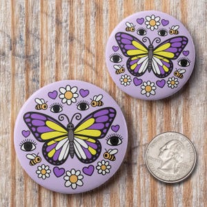 K&ouml;nnte beinhalten: Zwei runde, lila Kn&ouml;pfe mit Schmetterling-, Bienen-, Augen-, Herz- und Blumenmotiven. Der Schmetterling hat lila und gelbe Fl&uuml;gel. Eine amerikanische Vierteldollarm&uuml;nze liegt neben den Kn&ouml;pfen zum Gr&ouml;&szlig;envergleich. Die Kn&ouml;pfe liegen auf einer Holzoberfl&auml;che.