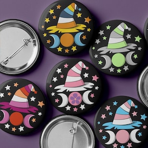 May include: A collection of black round buttons featuring colorful witch hats, crescent moons, and stars. The hats display various pride flag color schemes. Some buttons have a pin back.