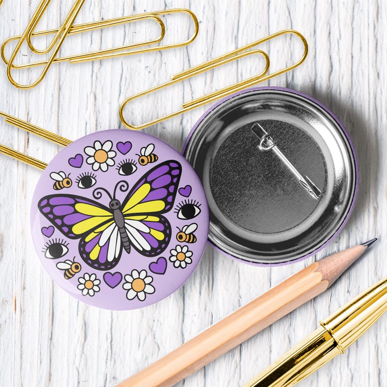 K&ouml;nnte beinhalten: Ein runder, lavendelfarbener Knopf mit einem Schmetterlingsdesign, mit lila und gelben Fl&uuml;geln, umgeben von Bienen, Augen, Blumen und Herzen. Die R&uuml;ckseite des Knopfes ist silberfarben. Goldene B&uuml;roklammern, ein Bleistift und ein Stift sind ebenfalls im Bild.