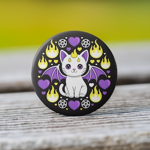 May include: A black button featuring a white cat with purple bat wings, horns, and a tail. The cat is surrounded by purple hearts, yellow flames, and pentagrams. The button is set against a blurred green and wooden background.