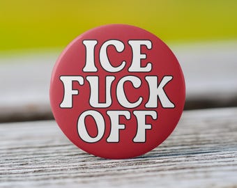 May include: A round, red button with the words "ICE FUCK OFF" in bold, white capital letters. The button is centered and set against a blurred background of green and yellow. The button is resting on a wooden surface.
