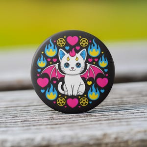 May include: A black button featuring a white cat with pink bat wings, a pink horn, and blue eyes. The cat is surrounded by pink hearts, blue and yellow flames, and gold pentagrams. The button is on a wooden surface.