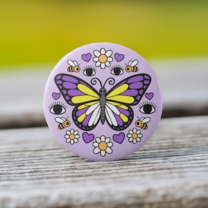 May include: A round, lavender button with a butterfly design. The butterfly has purple and yellow wings. The button also features bees, hearts, flowers, and eyes. The button is on a wooden surface.
