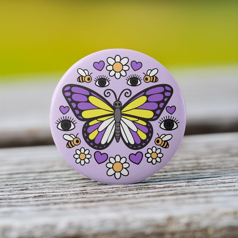 K&ouml;nnte beinhalten: Ein runder, lavendelfarbener Knopf mit einem Schmetterlingsdesign. Der Schmetterling hat lila und gelbe Fl&uuml;gel. Der Knopf zeigt auch Bienen, Herzen, Blumen und Augen. Der Knopf liegt auf einer Holzoberfl&auml;che.