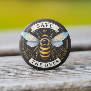 May include: Round black button with a detailed illustration of a bee, featuring yellow and black stripes, blue wings, and the text "SAVE THE BEES" on a white banner. The button has a decorative gold border.