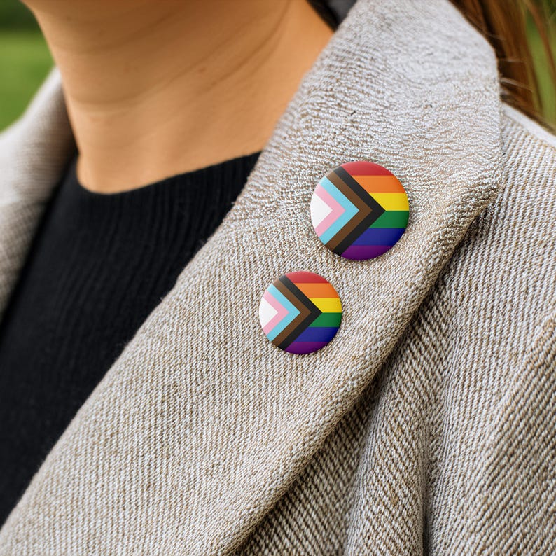 K&ouml;nnte beinhalten: Zwei runde Anstecknadeln mit dem Progress Pride Flaggen-Design sind auf einem hellbraunen Blazer befestigt. Die Anstecknadeln zeigen die Regenbogenflagge, die Farben der Transgender-Flagge und die Farben, die marginalisierte Farb-Communities repr&auml;sentieren.