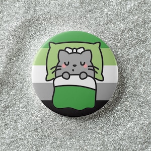 May include: A round button with an AroAce pride flag design. The button features a cartoon cat sleeping on a pillow under a green blanket. The background is a glittery silver.