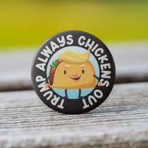May include: A black round button with the text "TRUMP ALWAYS CHICKENS OUT" encircling a cartoon taco with blonde hair. The taco has a smiling face and is wearing a blue outfit. The background is blurred.