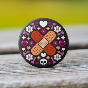 May include: A round, black button with a decorative design. The design features two crossed bandages in orange and tan, surrounded by flowers, hearts, and a skull. The button is set against a blurred green and wood background.