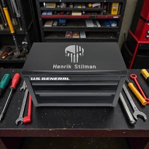 May include: A black US General tool chest with three drawers. The top of the chest features a skull design with an American flag motif and the name "Henrik Stilman". Tools are visible in the background.