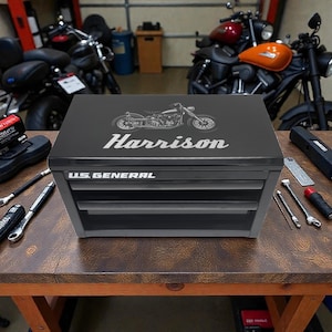 Peut inclure: Coffre à outils noir US General avec trois tiroirs, un motif de moto et le nom "Harrison" en argent. Le coffre est posé sur un établi en bois, avec des outils et des motos en arrière-plan.
