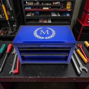 May include: A blue US General tool chest with drawers, featuring a silver initial "M" inside a laurel wreath on the lid. The chest is surrounded by tools, including screwdrivers and wrenches, on a dark surface.