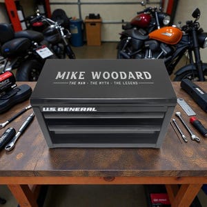 May include: A black and gray US General tool chest with three drawers, personalized with the name "MIKE WOODARD" and the tagline "THE MAN - THE MYTH - THE LEGEND" in silver lettering. Tools are scattered around the chest on a wooden surface.
