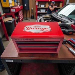 May include: A red US General tool chest with drawers, featuring the text "JERED'S Garage" in a white and red design on the top. The chest is sitting on a brown table in a garage setting.