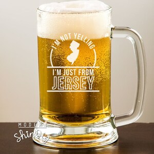 以下が含まれることがあります： 黄金色のビールと白い泡が入った透明なガラスのジョッキ。ジョッキには取っ手があり、「I'M NOT YELLING, I'M JUST FROM JERSEY」という白文字と、ニュージャージー州の白いアウトラインが描かれています。