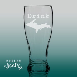 以下が含まれることがあります： 「Drink」の文字とミシガン州アッパー半島地図が刻まれた透明なガラスのビールグラス。グラスは曲線的な形状で、広い開口部があります。「Modern ShinDig」のロゴが下部にあります。