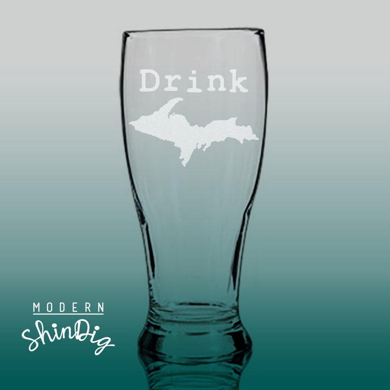 以下が含まれることがあります： 「Drink」の文字とミシガン州アッパー半島地図が刻まれた透明なガラスのビールグラス。グラスは曲線的な形状で、広い開口部があります。「Modern ShinDig」のロゴが下部にあります。