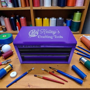 Peut inclure: Un coffre à outils de bricolage violet US General avec le texte "Kelley's Crafting Tools" sur le couvercle. Des pinceaux de différentes tailles et couleurs sont disposés autour du coffre, ainsi que des bobines de fil.