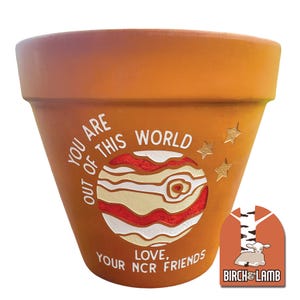 May include: A terracotta plant pot featuring a painted graphic of a planet with red and cream bands. The text reads "YOU ARE OUT OF THIS WORLD" and "LOVE, YOUR NCR FRIENDS" in white, with two gold stars. A logo with a lamb and "BIRCH & LAMB" is visible.