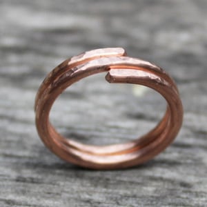 Puede incluir: Un anillo de cobre hecho a mano con una textura martillada. El anillo presenta un diseño abierto único con bandas superpuestas. El cálido metal marrón rojizo se muestra sobre un fondo neutro, destacando la simple elegancia del anillo.