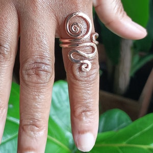 May include: A copper-colored ring with a hammered texture and a spiral design. The ring wraps around the finger with an open design, featuring a swirling top and a wavy lower section. The jewelry is displayed on a finger.