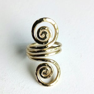 May include: A gold-colored ring with a hammered texture. The ring features a spiral design at the top and bottom, connected by multiple bands. The ring is set against a white background.