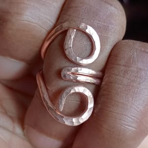Op de afbeelding: Een koperkleurige metalen ring met een bypass-ontwerp met twee open, cirkelvormige uiteinden. Het oppervlak van het metaal heeft een duidelijke gehamerde textuur, waardoor een reeks kleine, reflecterende inkepingen over de ringband ontstaat.
