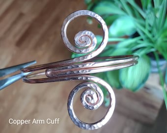 May include: Copper arm cuff with a hammered texture and spiral design. The cuff features two large spirals and a curved band. The text "Copper Arm Cuff" is visible at the bottom of the image.