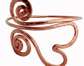 May include: Copper arm cuff with hammered texture and a spiral design. The cuff features a curved shape with two decorative spiral elements. The metal has a warm, reddish-brown color. This arm cuff is a piece of jewelry.