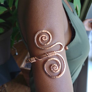 May include: Copper arm cuff featuring two spiral designs connected by wrapped wire. The hammered texture of the copper adds visual interest. The cuff is worn on the upper arm, showcasing its intricate design and craftsmanship.