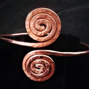 Può includere: Bracciale in rame con due spirali martellate. Il bracciale è di un caldo color marrone-rossiccio e ha una superficie strutturata. Le spirali sono centrate sul bracciale, creando un design unico.