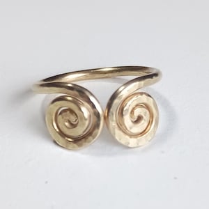 May include: A gold-colored ring with a hammered texture. The ring features two spiral designs on either side, connected by a smooth band. The ring is set against a white background.