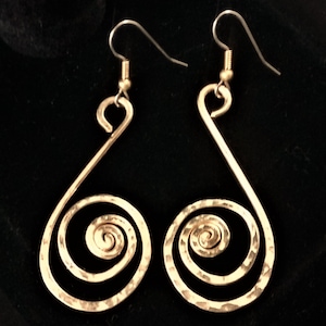 May include: A pair of gold-colored, hammered metal earrings. Each earring features a teardrop shape with a spiral at the bottom and a hook at the top. The earrings are displayed against a dark background.