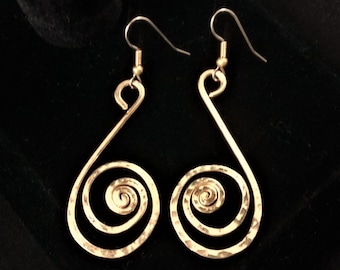 May include: A pair of gold-colored, hammered metal earrings. Each earring features a teardrop shape with a spiral at the bottom and a hook at the top. The earrings are displayed against a dark background.