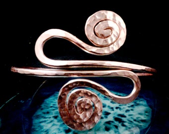 May include: Copper-colored arm cuff with a hammered texture. The cuff features a flowing design with two spiral accents, one at the top and one at the bottom. The metal has a warm, reddish-brown hue, and the design is set against a dark background.