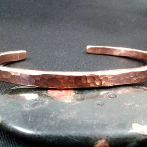 May include: A hammered copper bracelet with a smooth, polished finish. The bracelet is open and has a slightly curved shape.