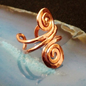 May include: A copper-colored ring with a hammered texture and a spiral design. The ring features two prominent spirals and a curved band, creating a unique and artistic look. The ring is displayed on a light blue surface.