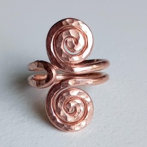 Puede incluir: Un anillo de cobre con un diseño de doble espiral y textura martillada. El metal tiene un acabado cálido de color marrón rojizo metálico. La banda es abierta, lo que permite ajustarlo, y las dos espirales están colocadas verticalmente.