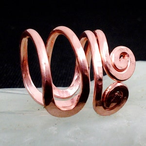 May include: A copper-colored ring with a unique spiral design. The ring features a continuous, looping form that wraps around, creating an open, airy feel. The metal has a hammered texture, adding visual interest. The ring's design is artistic and eye-catching.