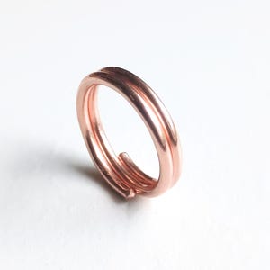 May include: A polished copper ring featuring a double-wrap design. The band is constructed from a single continuous piece of smooth, metallic copper wire that circles around twice, creating a layered appearance with rounded, finished ends.