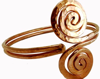 May include: Copper arm cuff with a hammered texture and spiral design. The cuff features a large, circular spiral element and a smaller spiral at the end, connected by two parallel bands. The metal has a warm, reddish-brown hue.